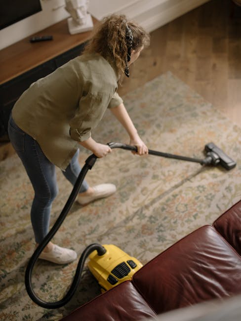 Close-up of a modern, upright vacuum cleaner with a transparent dust container filled with debris, positioned on a beige carpet in a residential room. The vacuum has a black and red body with a flexib
