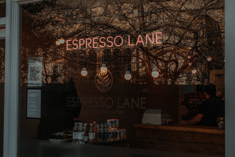 A view through the window of Espresso Lane, a coffee shop featuring a neon sign displaying the name 'ESPRESSO LANE' in pink that is illuminated against a backdrop of hanging light bulbs and a decorati
